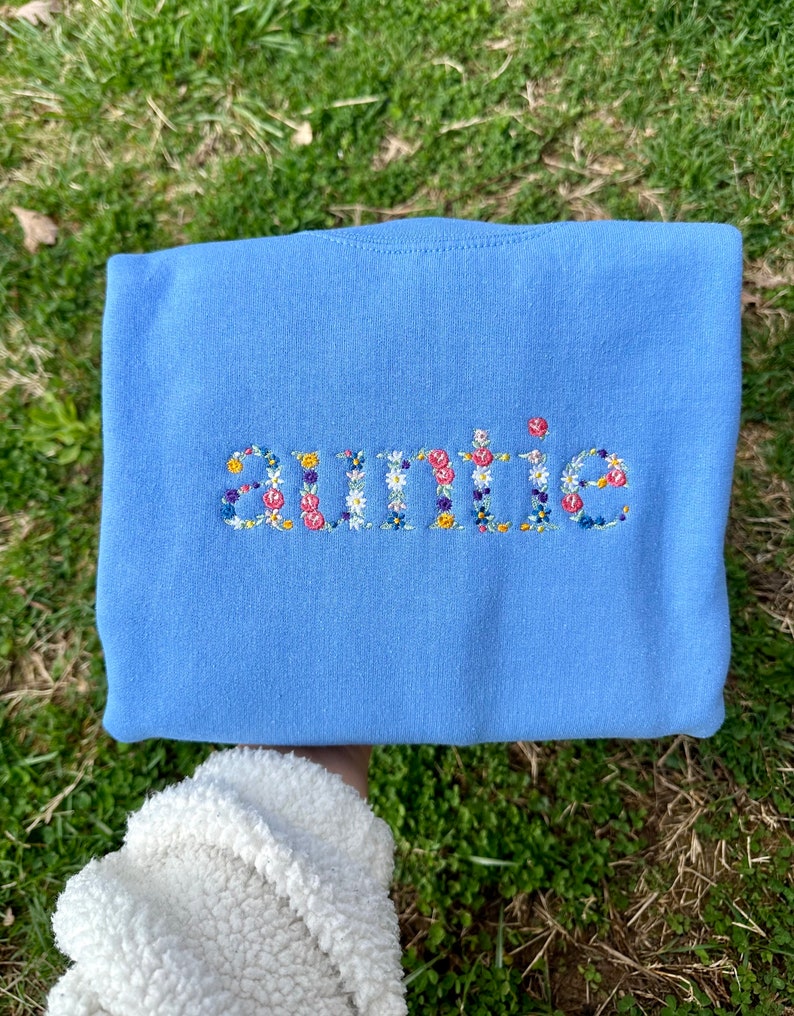 K&ouml;nnte beinhalten: Ein hellblauer Pullover mit dem Wort "auntie", das mit bunten Blumen bestickt ist.