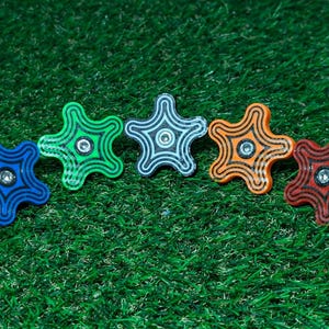Pode incluir: Cinco botões em forma de estrela dispostos em fila, cada um com uma cor distinta incluindo azul, verde, branco, laranja e vermelho. Cada botão apresenta um padrão de linhas pretas concêntricas e um parafuso central metálico.
