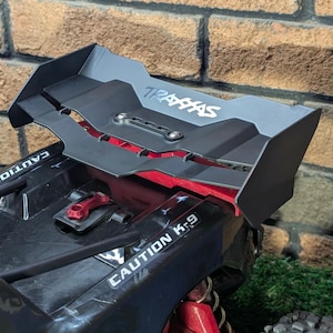 May include: A black remote-controlled vehicle spoiler featuring the Traxxas logo in white text. The spoiler is mounted on a black chassis with red accents and the text Caution K-9 printed on the side. The structure is made of matte black plastic.
