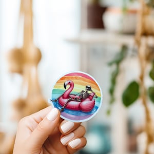 May include: A round sticker with a skeleton sitting on a pink flamingo float in a pool of water. The background is a rainbow with a sunset.