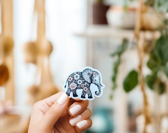 May include: A blue sticker with a black and white illustration of an elephant covered in floral designs. The elephant is facing right and has a trunk that is curled up.