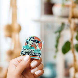 May include: A sticker featuring a vintage illustration of a woman using binoculars with the text "Oh, look! Nobody gives a shit."