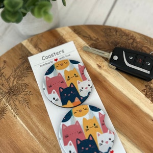 May include: Two round coasters with a colorful cat print design. The coasters are handmade with love and feature a variety of cats in different colors and poses. The text "Coasters handmade with love" is printed on a white card behind the coasters.