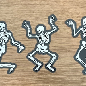 May include: Three black and white skeleton stickers with a dancing pose. The skeletons are outlined in black and have white bones.