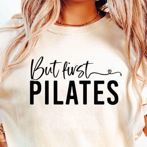 May include: A cream-coloured sweatshirt featuring the text "But first PILATES" in black. "But first" is in cursive, with a small heart. The word PILATES is in bold, block letters. The shirt has long sleeves.