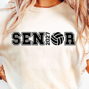 Op de afbeelding: Een crèmekleurig T-shirt met korte mouwen en zwarte blokletters met de tekst SENIOR, waarbij het getal 2027 en een volleybalafbeelding de letter I vervangen. Het ontwerp is gecentreerd op de borst in een vetgedrukt, collegiaal lettertype.