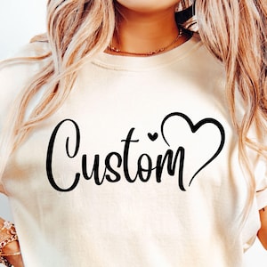 May include: Cream-colored t-shirt with the word "Custom" in a black script font, accompanied by a heart symbol. The shirt has short sleeves and a relaxed fit. The design is centered on the front of the shirt.