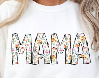 Può includere: Felpa bianca con la parola "MAMA" stampata in lettere nere bordate. Le lettere sono riempite con un motivo floreale di fiori rosa, arancioni e gialli.