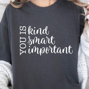 May include: Dark gray t-shirt with the words "YOU IS kind smart important" in white script. The shirt is a crew neck style and appears to be made of a soft material. The overall aesthetic is casual and comfortable.