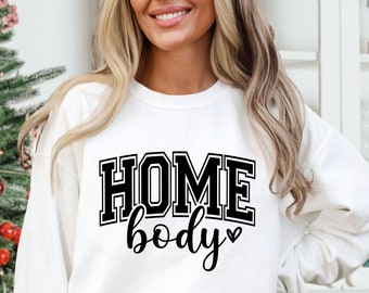 Può includere: Felpa bianca con testo nero che recita "HOMEbody" con un piccolo simbolo di cuore.