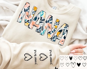 May include: A beige sweatshirt with the word "NANA" in large, stylized letters. The letters are filled with a floral pattern in shades of pink, yellow, and blue. The sweatshirt has two heart-shaped designs on the sleeve with the text "your text" next to each heart. There are also multiple heart-shaped designs in black and white on a white background.