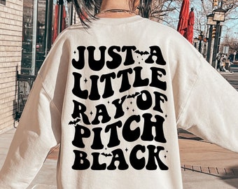 Puede incluir: Una sudadera blanca con el texto "Just a little ray of pitch black" en letras negras de estilo retro. El texto está rodeado de pequeños murciélagos y estrellas.