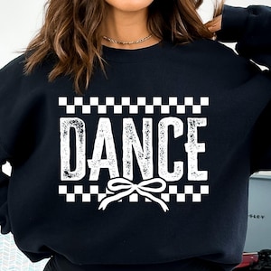Può includere: Felpa nera con un bordo a scacchi bianchi e la parola "DANCE" in un font usurato. Un fiocco bianco è centrato sotto la parola.