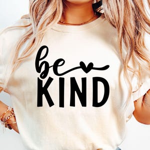 May include: A cream-coloured t-shirt featuring the words "be KIND" in black script and block letters. The word "be" is in cursive, with a heart symbol replacing the "o". The shirt has short sleeves.