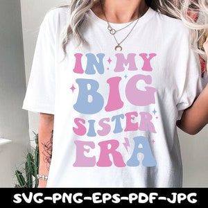May include: White t-shirt with the text "In My Big Sister Era" in pink and blue letters with stars and lightning bolts.