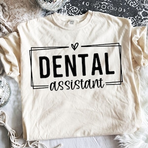 May include: A cream-coloured t-shirt featuring the words "DENTAL ASSISTANT" in black, within a black rectangular frame, with a small heart above "DENTAL". The shirt is made of a soft material.