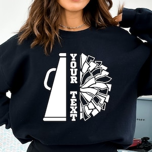 Pu&ograve; includere: Felpa nera con design di megafono e pompon da cheerleader bianchi. Il testo "YOUR TEXT" &egrave; stampato verticalmente. Il design &egrave; in stile grafico audace, ideale per le squadre di cheerleader.