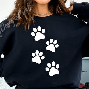 Op de afbeelding: Zwart sweatshirt met een wit poot afdruk ontwerp. De poot afdrukken zijn in het midden van het sweatshirt in een cluster gerangschikt.