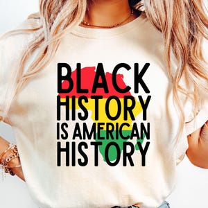 Puede incluir: Una camiseta blanca con el texto "BLACK HISTORY IS AMERICAN HISTORY" en letras negras. El texto se superpone a un gráfico del continente africano en rojo, amarillo y verde.