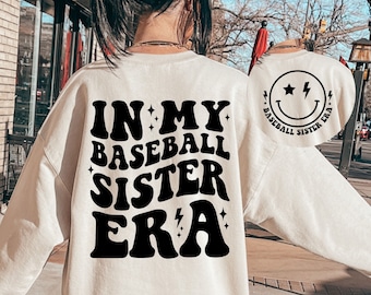 May include: A white sweatshirt with the text "IN MY BASEBALL SISTER ERA" in black retro font. The back of the sweatshirt has a black smiley face with lightning bolt and stars, and the text "BASEBALL SISTER ERA".