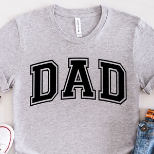 Op de afbeelding: Een grijs T-shirt met het woord "DAD" in zwarte blokletters.