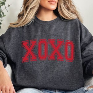 May include: Dark gray sweatshirt with the letters "XOXO" in red, outlined in a darker red. The sweatshirt is a crew neck style, and the letters have a distressed, vintage look.