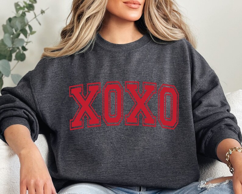 May include: Dark gray sweatshirt with the letters "XOXO" in red, outlined in a darker red. The sweatshirt is a crew neck style, and the letters have a distressed, vintage look.