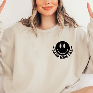 Pu&ograve; includere: Una felpa beige con un'immagine di un viso sorridente nero e il testo "Band Mom Era" sotto.