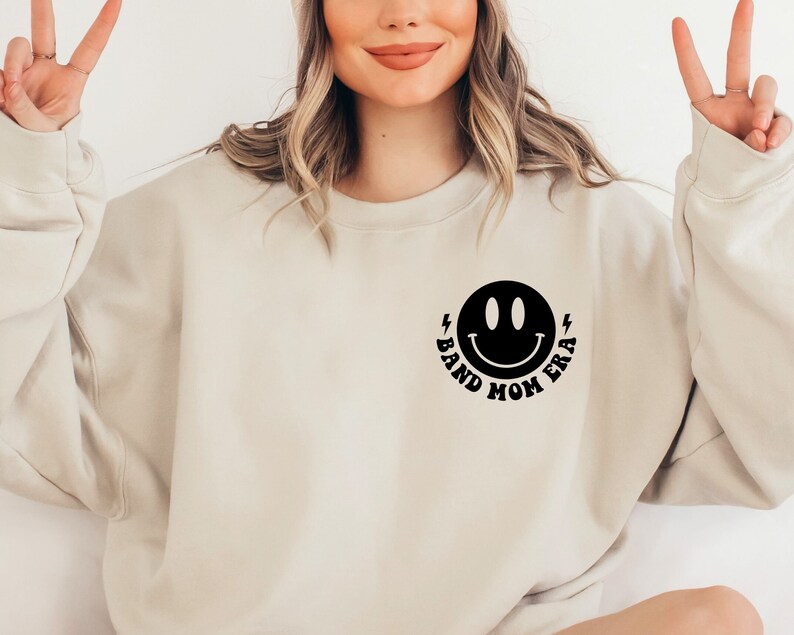 Pu&ograve; includere: Una felpa beige con un'immagine di un viso sorridente nero e il testo "Band Mom Era" sotto.