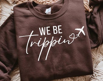 Può includere: Una felpa girocollo marrone scuro con la scritta bianca "WE BE" sopra la parola "trippin" in carattere corsivo, seguita da una piccola icona a forma di cuore e dal disegno di un aereo che vola verso il lato destro del capo.