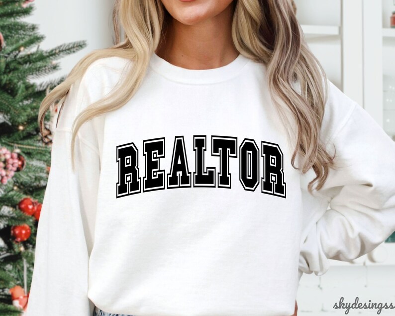May include: White sweatshirt with black text that reads "REALTOR" in a varsity style font.