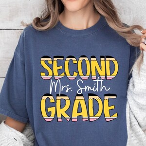 May include: A navy blue t-shirt with the text "Second Grade" in yellow and pink lettering with a pencil design. The text "Mrs. Smith" is in white lettering.
