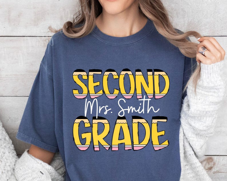 May include: A navy blue t-shirt with the text "Second Grade" in yellow and pink lettering with a pencil design. The text "Mrs. Smith" is in white lettering.