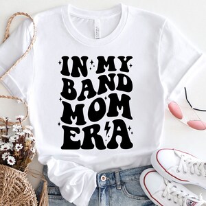 Pu&ograve; includere: Maglietta bianca con testo nero che recita "In My Band Mom Era" in uno stile di font retr&ograve;. La maglietta &egrave; abbinata a jeans blu e scarpe da ginnastica bianche.