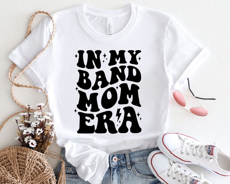 Pu&ograve; includere: Maglietta bianca con testo nero che recita "In My Band Mom Era" in uno stile di font retr&ograve;. La maglietta &egrave; abbinata a jeans blu e scarpe da ginnastica bianche.