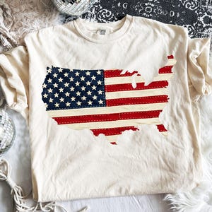 Könnte beinhalten: Cremefarbenes T-Shirt mit einem aufgenähten Design der amerikanischen Flagge in Rot, Weiß und Blau. Die Flagge hat die Form der Landkarte der Vereinigten Staaten. Das Shirt hat kurze Ärmel und einen Rundhalsausschnitt.