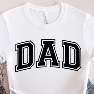 Op de afbeelding: Wit T-shirt met een zwarte "DAD"-afbeelding in een varsity-letter-stijl.