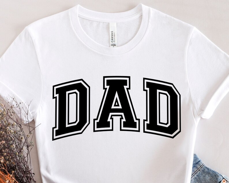 Op de afbeelding: Wit T-shirt met een zwarte "DAD"-afbeelding in een varsity-letter-stijl.