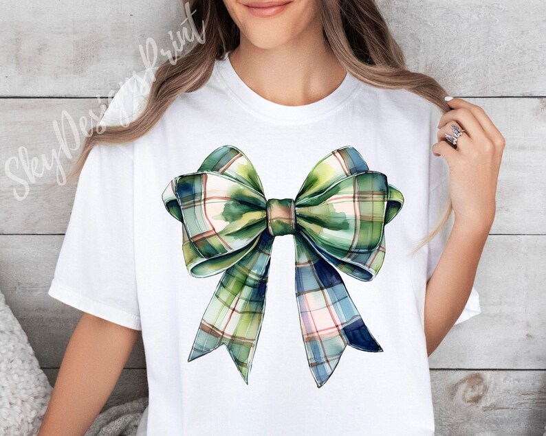 Peut inclure: T-shirt blanc avec un grand n&oelig;ud aquarelle. Le n&oelig;ud pr&eacute;sente un motif &agrave; carreaux dans des tons de vert, de bleu et de rose. Le design est centr&eacute; sur le t-shirt, cr&eacute;ant un look simple mais accrocheur.