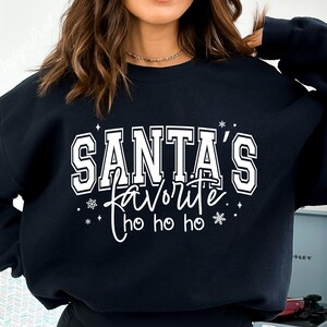 May include: Black sweatshirt with the text "SANTA'S FAVORITE" in white block letters, with "favorite" in a cursive font. The design includes snowflakes and "ho ho ho". A festive holiday-themed top.