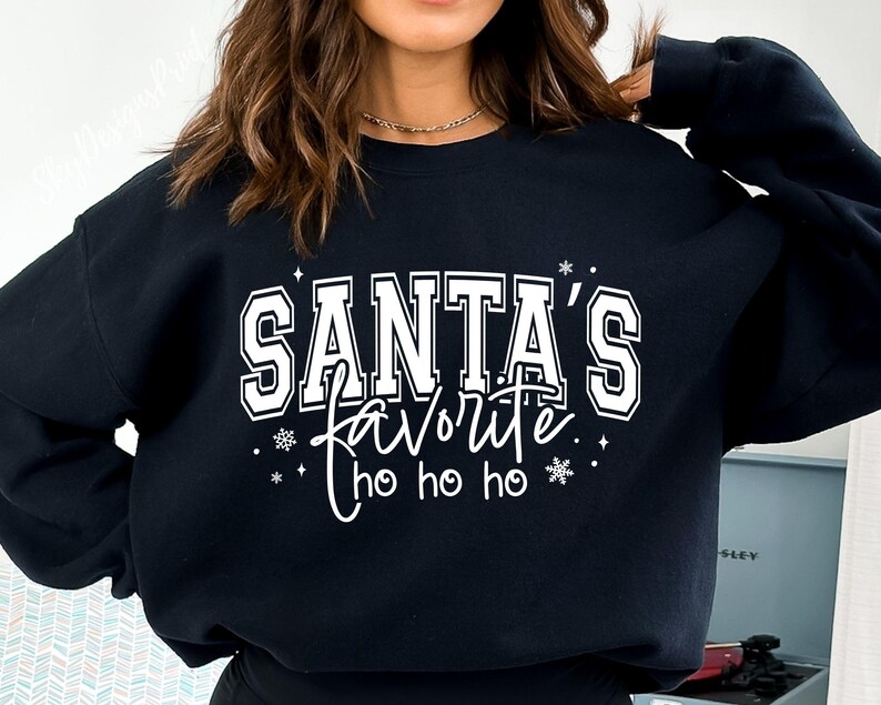 May include: Black sweatshirt with the text "SANTA'S FAVORITE" in white block letters, with "favorite" in a cursive font. The design includes snowflakes and "ho ho ho". A festive holiday-themed top.