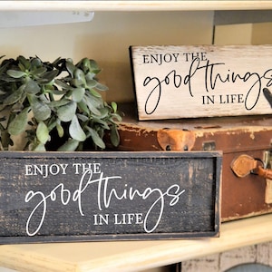 May include: Two rectangular wooden signs with the phrase "Enjoy the good things in life" in cursive. One sign is black, the other is white. A small black heart is on the white sign. A potted plant and a vintage suitcase are also in the image.