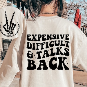 May include: A cream colored sweatshirt with a black graphic on the back that says "Expensive Difficult & Talks Back" in a retro font. A smaller graphic of a skeleton hand making a peace sign is on the left sleeve.