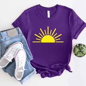 May include: Purple t-shirt with a yellow sun graphic. The sun has rays coming out of it. The t-shirt is on a white surface with a pair of blue jeans and white sneakers.