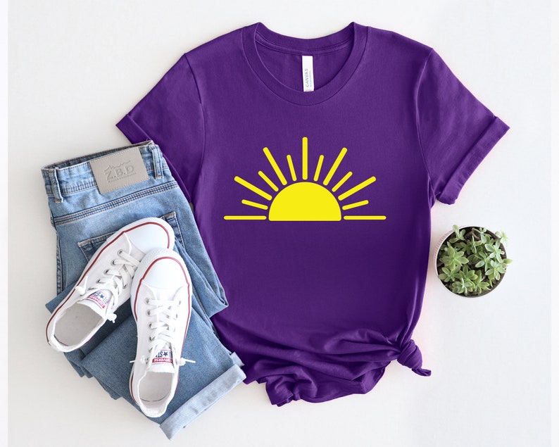 May include: Purple t-shirt with a yellow sun graphic. The sun has rays coming out of it. The t-shirt is on a white surface with a pair of blue jeans and white sneakers.