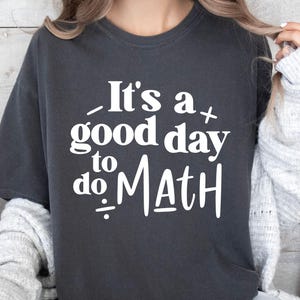 May include: Dark gray t-shirt with the white text "It's a good day to do MATH". The shirt has a crew neck and short sleeves. The text includes mathematical symbols.