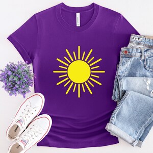 May include: Purple t-shirt with a yellow sun graphic. The shirt is laid flat on a white surface with a pair of white sneakers and a pair of blue jeans.