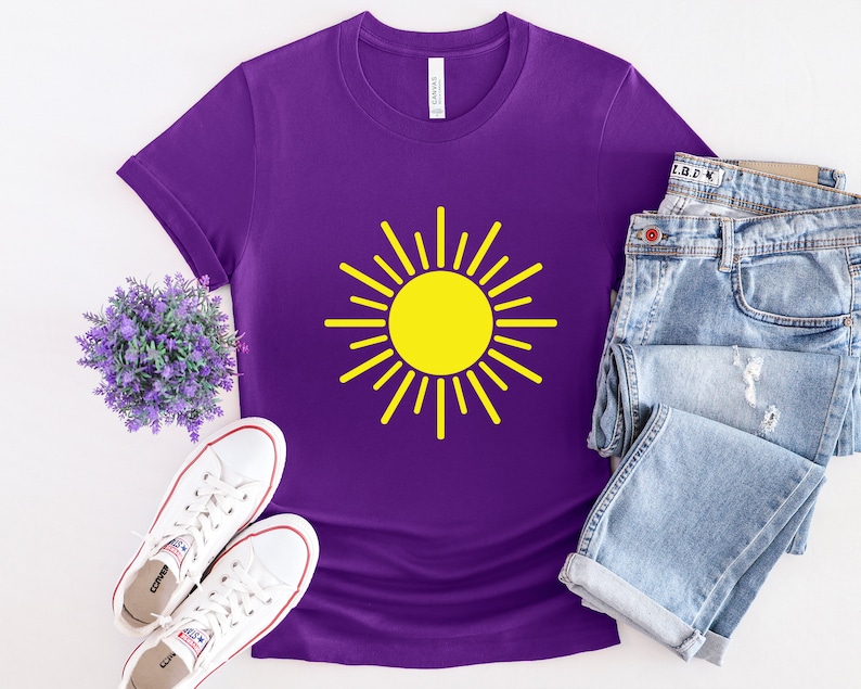 May include: Purple t-shirt with a yellow sun graphic. The shirt is laid flat on a white surface with a pair of white sneakers and a pair of blue jeans.