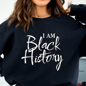 Puede incluir: Sudadera negra con el texto "I am Black History" en letras blancas.