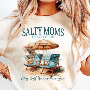May include: A white t-shirt with a graphic design of two beach chairs under a beach umbrella with palm trees. The text "Salty Moms Beach Club" and "Girls Just Wanna Have Sun" are printed on the shirt.